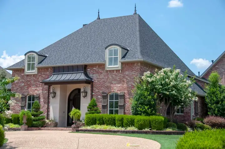 Drone view of a brick French-country home with a steep, finished dark shingle roof and dormers